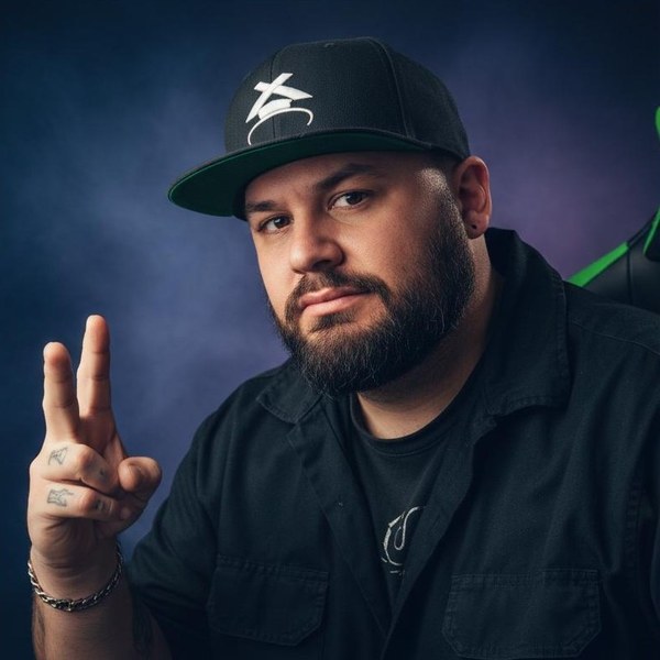 Monakai portrait photo - bearded electronic music producer wearing a black baseball cap with white logo, black shirt, and silver chain bracelet, making a peace sign gesture against a dark blue gradient background